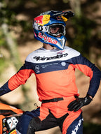 Alpinestars Pro-Dura Jersey, motorcross shirt in Navy, Hot Orange en wit, voorzien van opvallende branding op de borst en laserperforaties, motorcrosser met Red Bull-racenhelm, professionele uitrusting voor enduro- en off-road-prestaties.