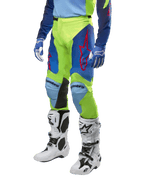 Alpinestars 2024 Racer Hoen broek, motorcrossbroek, fluogeel en donkerblauw, voorzien van rode sterlogo's en zwarte panelen aan de binnenkant van de knieën, gedragen met een bijpassende jersey en witte motorcrosslaarzen, technische uitrusting voor off-road racen