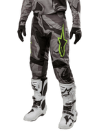 Alpinestars 2024 Racer Tactical broek, motorcrossbroek, grijs en zwart camouflagepatroon, limoengroen sterlogo, gedragen met witte en zwarte laarzen en zwarte handschoenen, off-road motoruitrusting