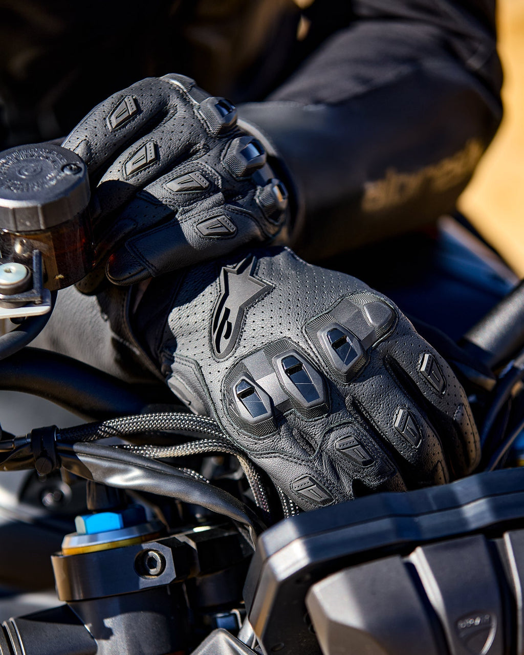 Alpinestars SP X 7 handschoenen, zwarte geperforeerde leren motorhandschoenen met TPU-knokkelbeschermers en vinger-sliders, afgebeeld op de handen van een motorrijder die een motorstuur vasthoudt
