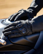 Alpinestars SP X 7 handschoenen, zwart, motorhandschoenen met korte manchet en geperforeerd leer, handen rustend op een donkere brandstoftank van een motorfiets, voorzien van zichtbare TPU-knokkelbescherming met dubbele dichtheid en logo in reliëf, ontworpen voor sport- en straatgebruik.