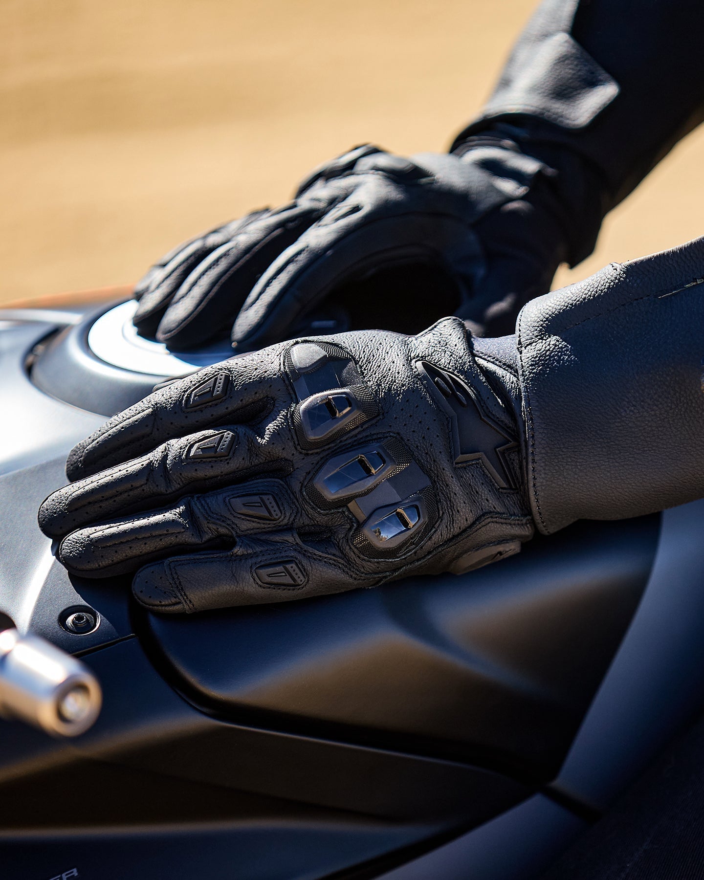 Alpinestars SP X 7 handschoenen, zwart, motorhandschoenen met korte manchet en geperforeerd leer, handen rustend op een donkere brandstoftank van een motorfiets, voorzien van zichtbare TPU-knokkelbescherming met dubbele dichtheid en logo in reliëf, ontworpen voor sport- en straatgebruik.
