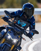 Motorrijder die in een bocht leunt met de Alpinestars T-Stunt Air Jacket in zwart, donkerblauw en helderblauw, gecombineerd met een zwarte helm en handschoenen, actiefoto op een verharde weg, sportieve motoruitrusting