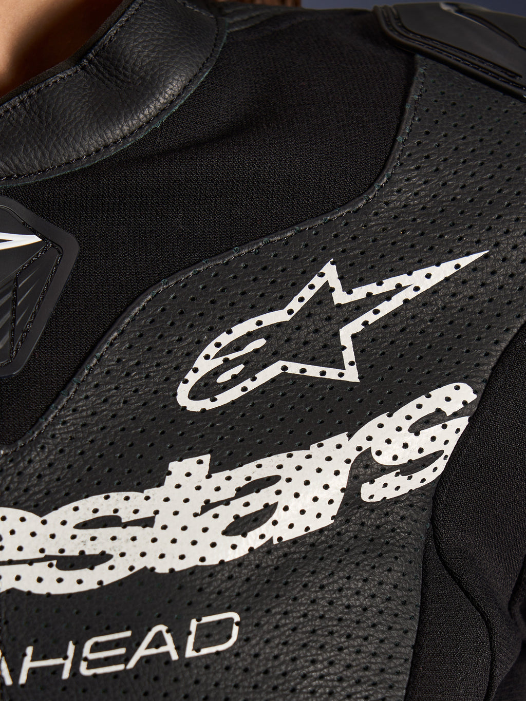 Close-up van het Alpinestars Stella GP-R7 2-delig lederen motorpak in het zwart, met geperforeerde lederen panelen, een groot wit Alpinestars-logo op de borst en ademende stretchstof gedeeltes.