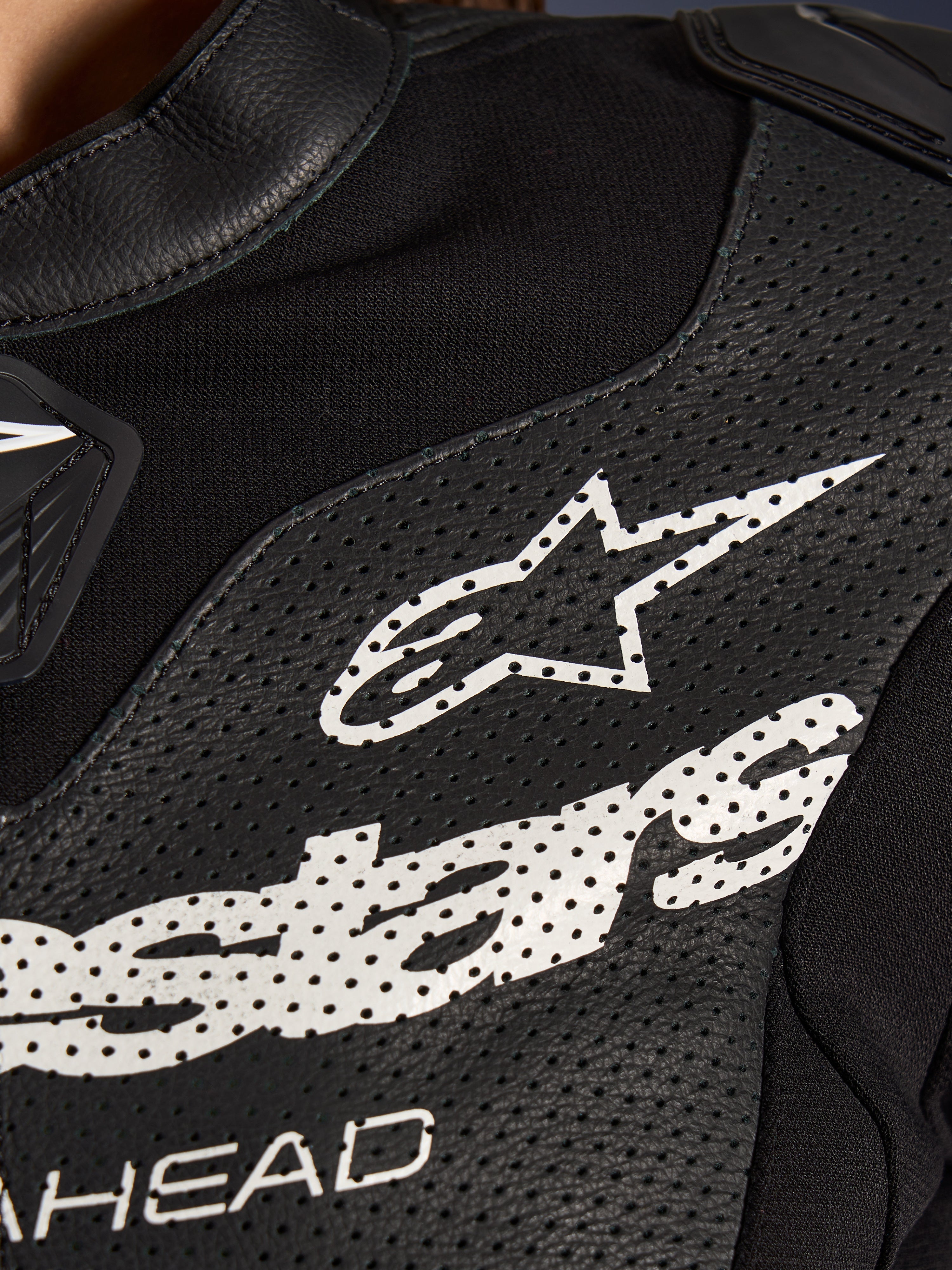 Close-up van het Alpinestars Stella GP-R7 2-delig lederen motorpak in het zwart, met geperforeerde lederen panelen, een groot wit Alpinestars-logo op de borst en ademende stretchstof gedeeltes.