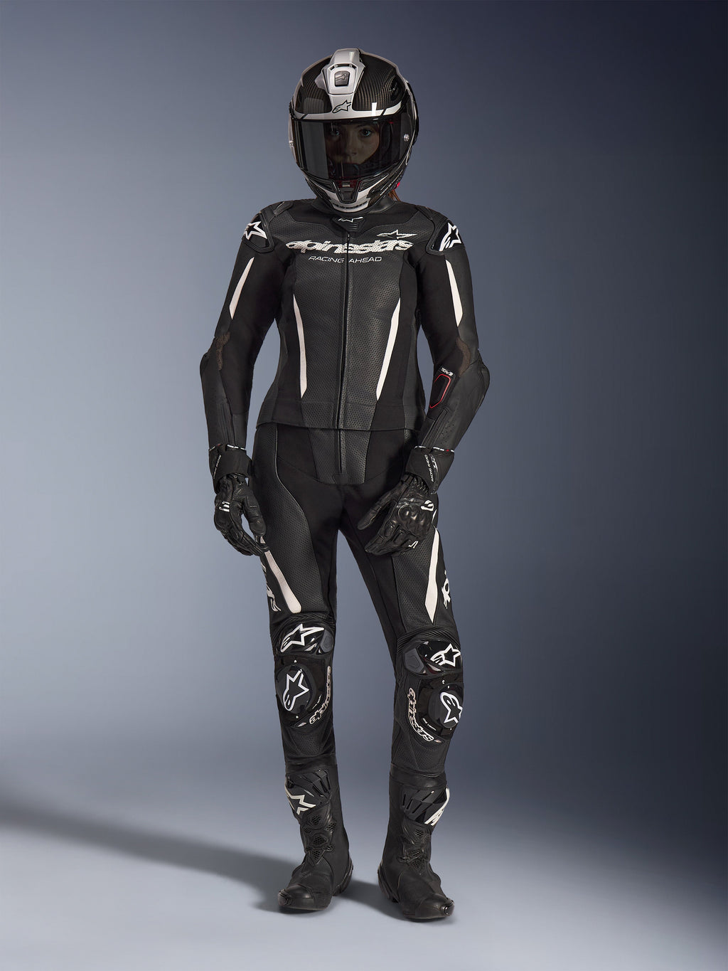 Vrouwelijke motorrijder in het Alpinestars Stella GP-R7 2-delig leren pak in zwart met witte accenten, compleet met een carbon fiber integraalhelm, zwarte racehandschoenen en laarzen, voorzien van geperforeerde leren panelen en geïntegreerde beschermende bepantsering, professionele motorrace-uitrusting voor circuit- en straatprestaties