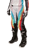Alpinestars Stella Techstar 2024 damesbroek, motorcrossbroek, zwart multicolor met diagonale rode, gele en blauwe strepen, gedragen met zwarte motorlaarzen en witte handschoenen, met beschermende zwarte kniepanelen en opvallende branding voor off-road racing.