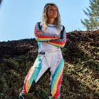 Vrouwelijke motorcrossrijder in Alpinestars Stella Techstar-broek 2024 voor dames en bijpassende jersey, Black Multicolor-ontwerp met witte basis en blauwe, gele en rode strepen, staand met de armen over elkaar op een crossbaan, professionele off-road outfit