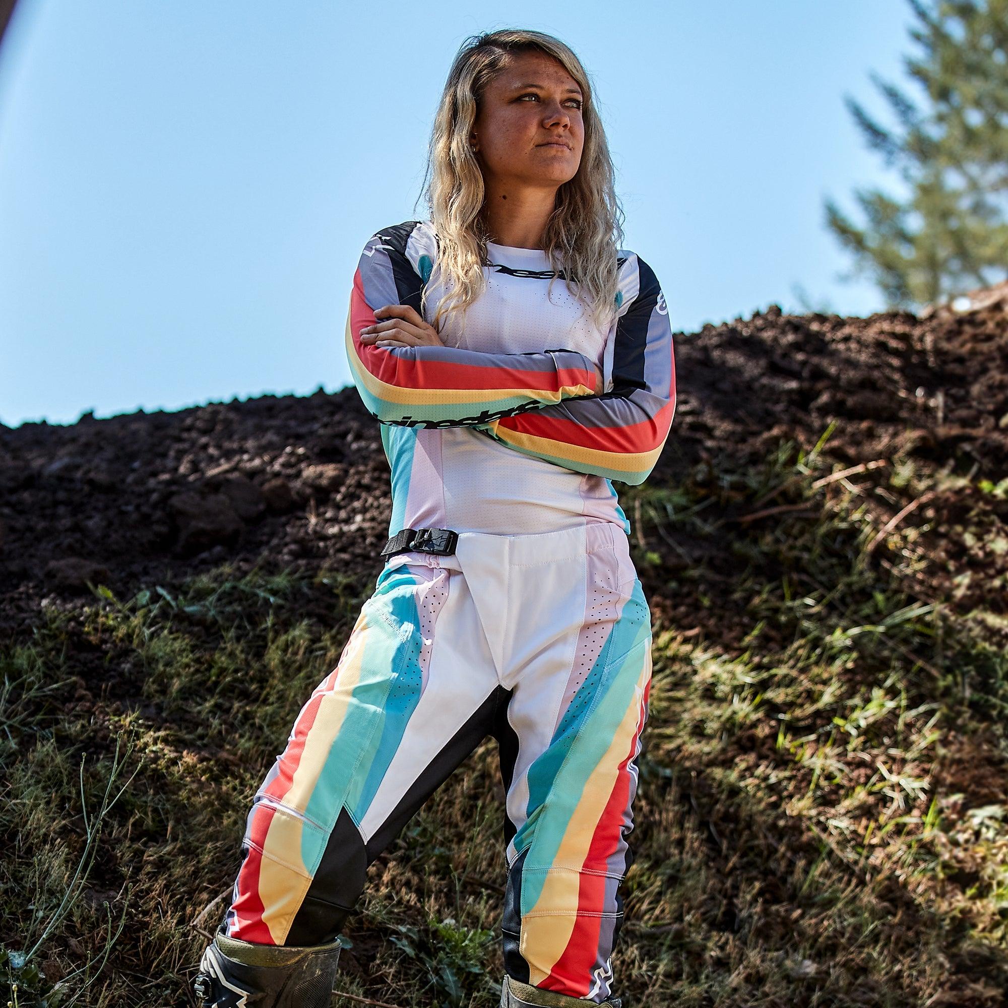 Vrouwelijke motorcrossrijder in Alpinestars Stella Techstar-broek 2024 voor dames en bijpassende jersey, Black Multicolor-ontwerp met witte basis en blauwe, gele en rode strepen, staand met de armen over elkaar op een crossbaan, professionele off-road outfit