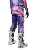 Alpinestars 2024 Techstar Ocuri crossbroek, motorcross broek, paars met feloranje accenten en grote logo-opdruk op het zitvlak, achteraanzicht, gedragen met witte motorcrosslaarzen en zwarte handschoenen