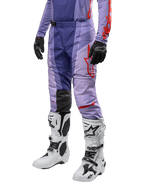 Alpinestars 2024 Techstar Ocuri crossbroek, motorcross broek in paars met fel oranje en rode accenten, rood logo op het bovenbeen, gedragen met witte motorcrosslaarzen en zwarte handschoenen, duurzame stretchconstructie voor off-road gebruik.
