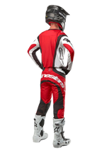 Alpinestars 2024 Techstar Ocuri broek, motorcrossbroek, Mars rood wit zwart, achteraanzicht van een motorcrosser in volledige race-uitrusting, inclusief bijpassende jersey, zwarte helm en wit-zwarte laarzen, met de nadruk op de Alpinestars-branding en duurzame materiaalconstructie.