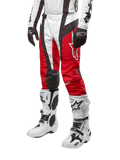 Alpinestars 2024 Techstar Ocuri crossbroek, motorcross broek, Mars rood, wit en zwart, gedragen met bijpassende wit-zwarte jersey, zwarte handschoenen en witte motorlaarzen met gespen, technische beschermende uitrusting op een witte achtergrond