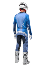 Alpinestars 2024 Techstar Pneuma broek, motorcrosser van achteren gezien in donkerblauw en lichtblauw, gecombineerd met bijpassende blauwe jersey, witte helm, handschoenen en laarzen, met opvallende Alpinestars-branding op het zitvlak en de mouwen.