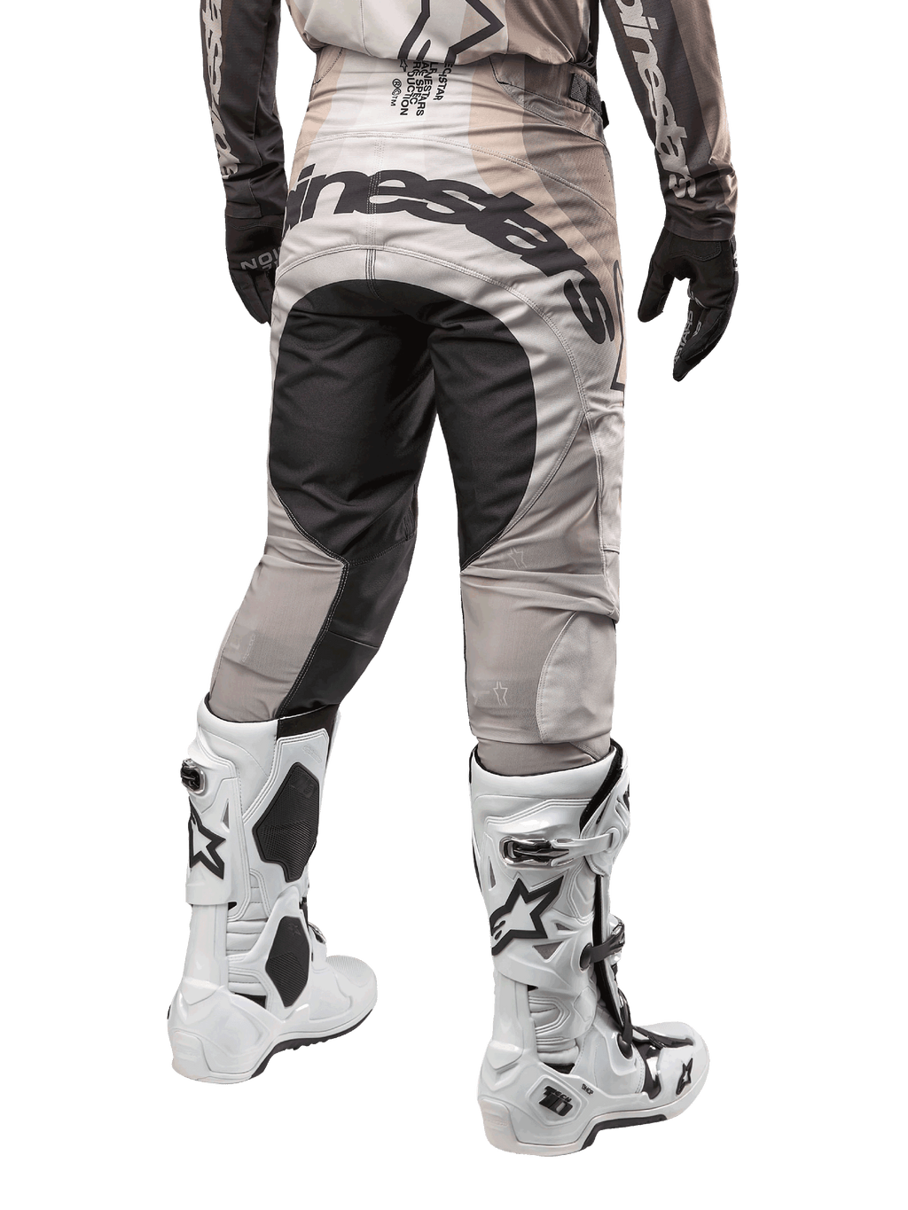 Alpinestars 2024 Techstar Pneuma crossbroek, motorcross broek, Dark Sand en Iron Dust Gray, achteraanzicht vanaf de taille naar beneden, opvallende zwarte Alpinestars-branding op het zitvlak, gedragen met wit-zwarte motorcrosslaarzen en zwarte handschoenen, duurzaam race-fit ontwerp voor off-road competitie