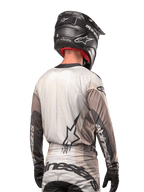Alpinestars 2024 Techstar Pneuma Jersey, motocross shirt rugaanzicht, kleur Dark Sand Iron Dust Gray, zwarte helm met carbon-afwerking, groot sterlogo op de rug en Alpinestars-branding op de mouwen