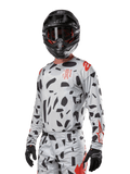 Alpinestars 2024 Techstar Rantera Jersey, motorcross shirt, Haze Gray Camo met zwarte abstracte patronen en rode accenten, ontwerp met lange mouwen gedragen met bijpassende zwarte helm en handschoenen, technische off-road race-uitrusting