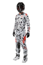 Alpinestars 2024 Techstar Rantera broek, motorcrossbroek, Haze Gray Camo, gedragen met bijpassende jersey, zwarte helm en witte en zwarte laarzen, met zwarte abstracte patronen en rode accenten, complete off-road race-uitrusting