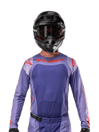 Alpinestars 2024 Techstar Ocuri Jersey, paars en feloranje, gedragen door een motorcrosser met een zwarte helm en crossbril, voorzien van lasergesneden perforaties op de borst, atletische racepasvorm en opvallende rode branding op de schouders en borst.