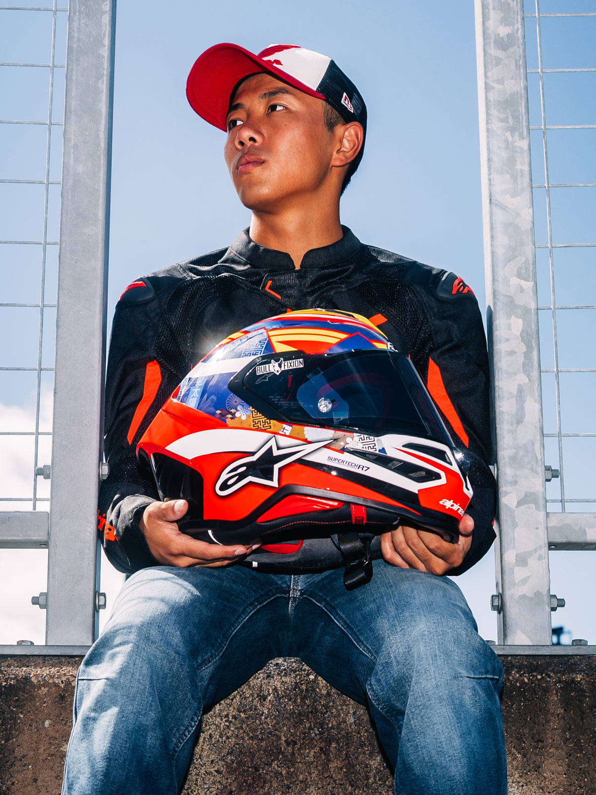 Alpinestars S-R7 Taiyo Furusato helm, integraal motorracehelm, rood, wit en blauw grafisch design, vastgehouden door coureur Taiyo Furusato in een zwarte jas, buiten langs het circuit.