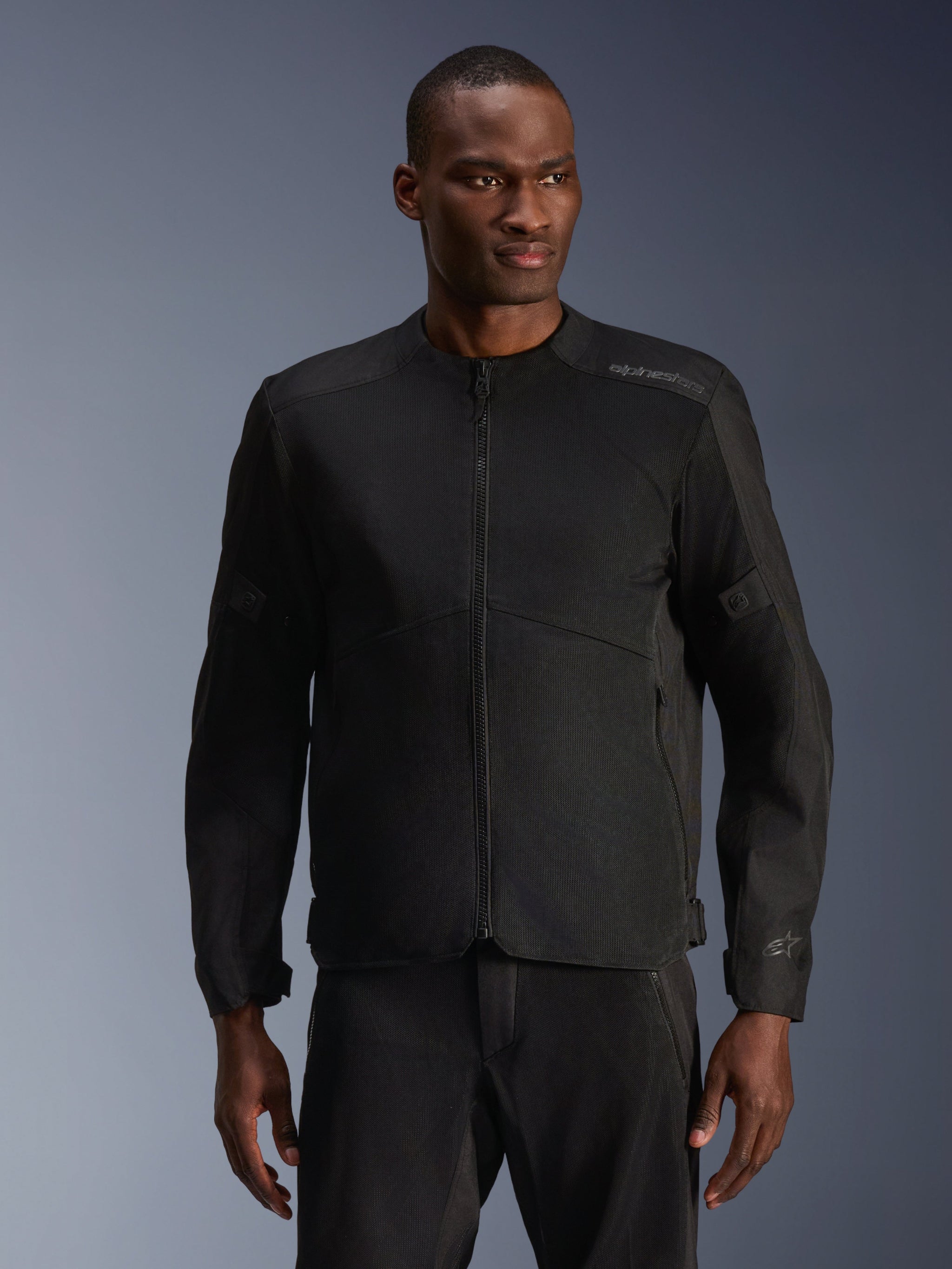 Mannelijk model draagt Alpinestars C-1 Air Jacket in het zwart, eigentijdse motorjas met uitgebreide tech mesh-panelen en een volledige ritssluiting aan de voorzijde, gecombineerd met bijpassende zwarte motorbroek, urban lifestyle en woon-werkverkeer uitstraling
