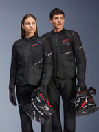 Man en vrouw dragen Alpinestars Andes Air Drystar®-jassen, zwarte adventure touring-kleding met mesh-panelen en reflecterende details, bijpassende zwarte broek en handschoenen, houden motorcrosshelmen vast, studio-opname van professionele motorkleding