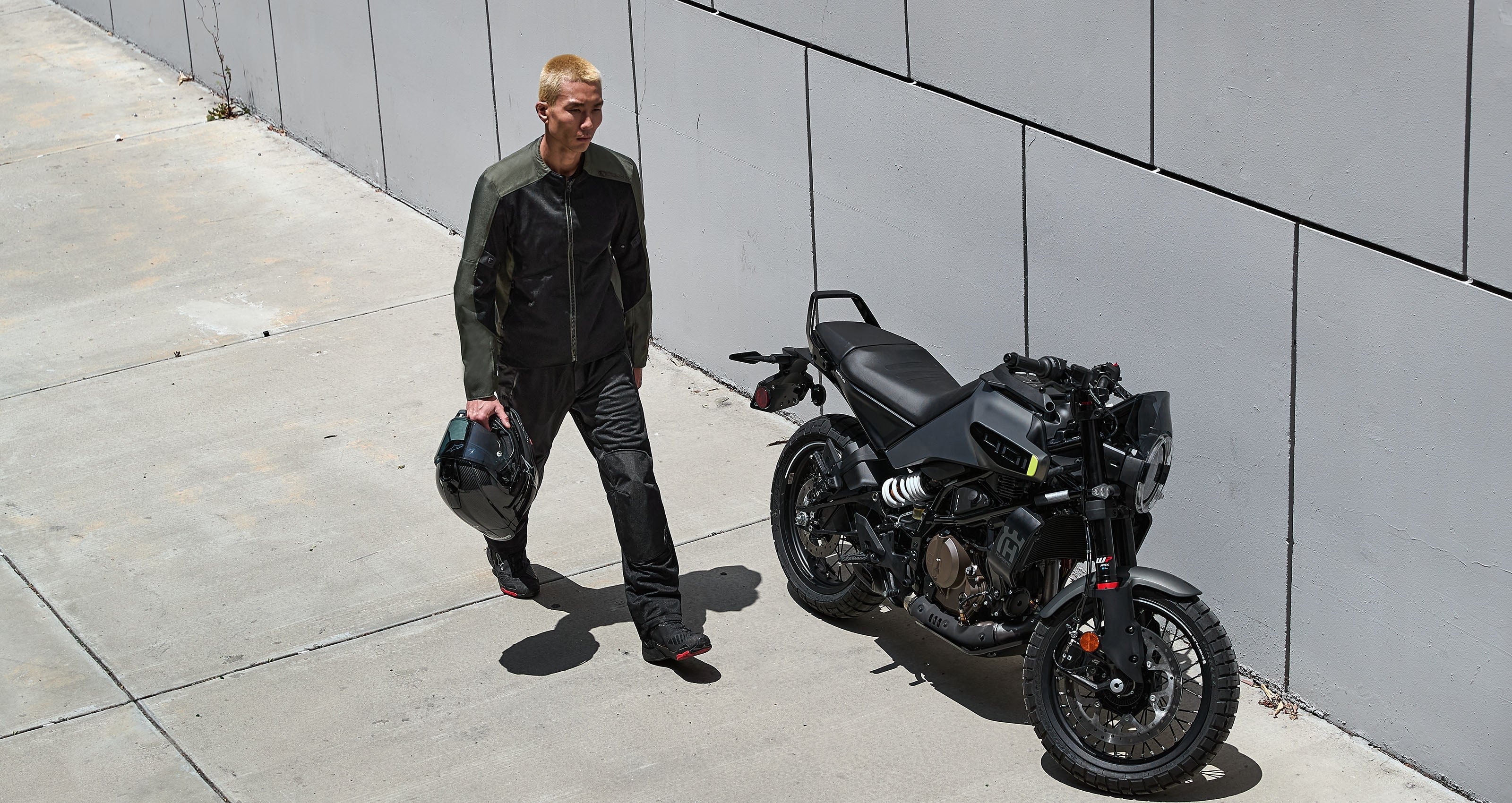 Alpinestars C-1 Air Jacket in black and olive green, urban motorcycle jacket worn by a man holding a helmet near a bike.